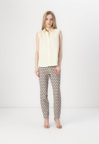 Blusa sin mangas de color amarillo claro con botones en el frente, combinada con pantalones ajustados que presentan un patrón geométrico en marrón y blanco.