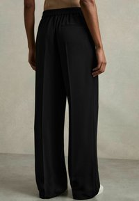 Persoon draagt losse zwarte broek met elastische tailleband, staat op blote voeten op een lichtgrijze vloer tegen een effen muur.