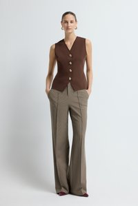 Gilet marrone senza maniche con scollatura a V e cinque bottoni dorati, abbinato a pantaloni a quadri a vita alta e gamba larga in tonalità attenuate.