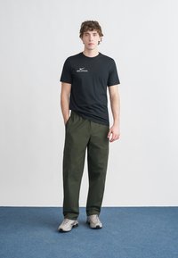 Crna Nike majica s logotipom, zelene cargo hlače i sivo tenisice. Ogrtač ima klasični kroj s vidljivim džepovima na hlačama.