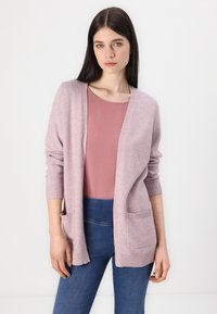 Cardigan rosa chiaro con una texture morbida, design aperto, polsini a costine e due tasche frontali, indossato sopra una maglia rosa e jeans blu.