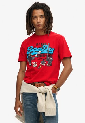 Camiseta de algodón roja con un estampado gráfico en azul y negro con elementos de dibujos animados. Combinada con unos vaqueros y un suéter beige atado a la cintura.