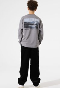 Grijze langwerpige shirt met een landschapsgrafiek op de achterkant, gecombineerd met zwarte corduroy broek. Witte sneakers maken de outfit compleet.