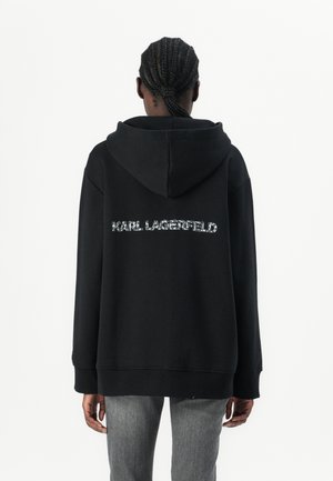Mustat ylisuuret hupparit, joissa on tekstuurinen "KARL LAGERFELD" -logo selässä, pitkät hihat ja etutasku, yhdistettyinä harmaisiin farkkuihin.