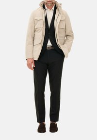Beige parka-jakke med hætte, flere frontlommer, parret med en sort dragt, cremet skjorte og brune ruskinds loafers.