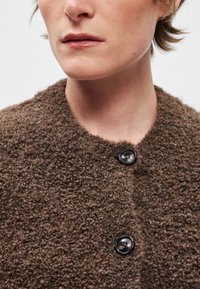 Cardigan de malha texturada castanha com decote redondo e dois botões pretos na parte da frente. Tecido suave com uma aparência peluda.