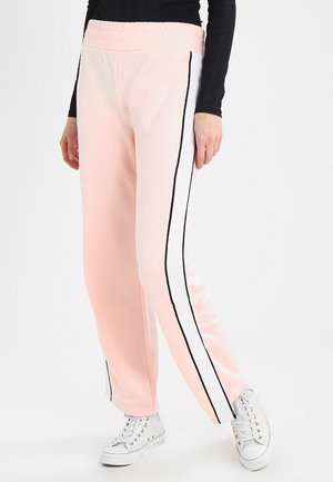 Kobieta w jasnoróżowych spodniach dresowych z białymi i czarnymi paskami po bokach oraz białych trampkach, stojąca na jednolitym tle.