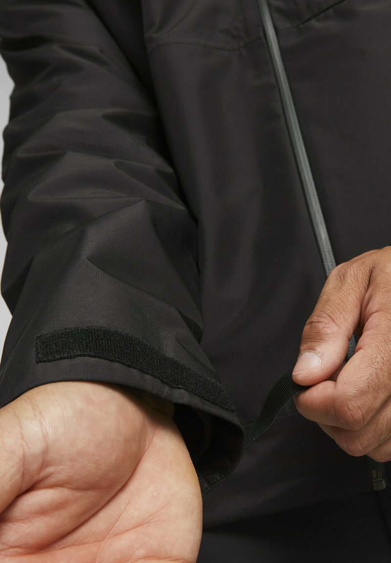 Schwarze wasserdichte Jacke mit Ärmel aus Velcro und elastischer Verstellung. Merkmale strukturierter Stoff mit glatter Oberfläche und grauem Reißverschluss.