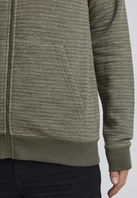 Veste à capuche zippée vert olive présentant un design texturé en côtes, une poche avant et des poignets ajustés. Tissu doux avec un motif rayé subtil.