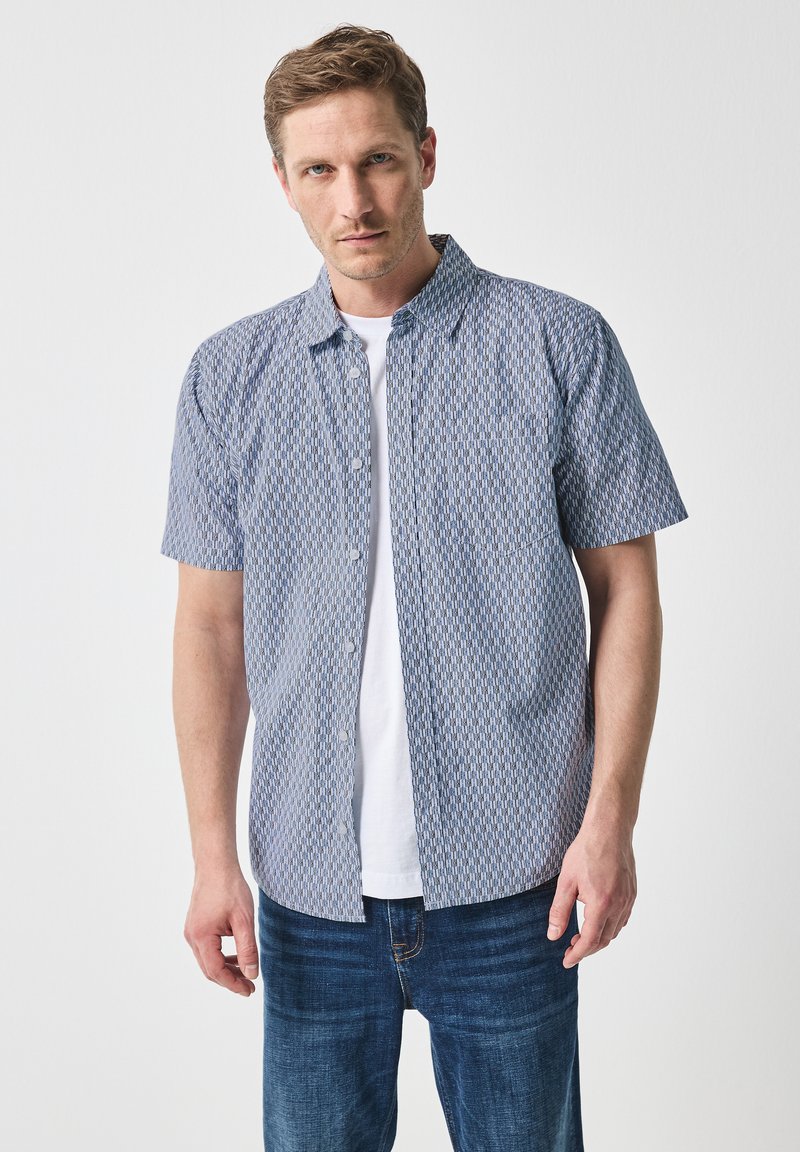 Košile s krátkým rukávem z modro-bílého vzorovaného materiálu, s límcem a rovnými lemy, nošená přes bílý tričko.