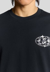 Juodi medvilniniai marškinėliai su apskritu kaklu. Ant krūtinės dalies matomas sidabrinis logotipo dizainas, išryškėjęs ant tekstūruoto audinio fono.