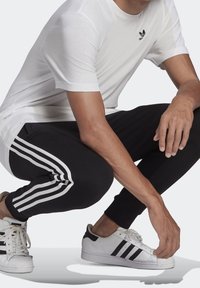 Maglietta bianca con logo Adidas nero, pantaloni jogger neri con strisce bianche e sneakers bianche con accenti neri, in posizione accovacciata.