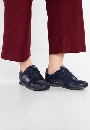 Osoba stojící na světlé podlaze ve tmavě modrých lesklých texturovaných teniskách se stejnobarevnými tkaničkami a s krátkými bordó kalhotami.