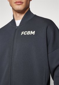 Dunkelgrau zip-up Bomberjacke aus strukturiertem Stoff, mit Stehkragen und gesticktem "FCBM"-Logo auf der Vorderseite.