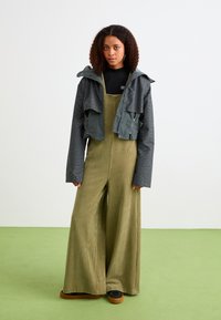Giacca corta e fantasia in verde scuro sovrapposta a una tuta lunga in corduroy oliva. L'outfit presenta pantaloni a gamba larga e calzature nere.