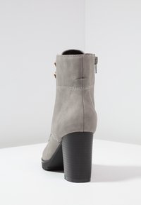 Bottine en daim gris avec talon carré empilé et fermeture éclair latérale, vue de dos sur une surface blanche avec un fond uni.