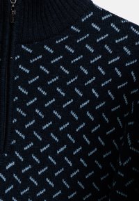 Gebreide trui met een hoge kraag in marineblauw, met een geometrisch patroon in lichtblauw. Het materiaal heeft een zachte textuur en een subtiele glans.