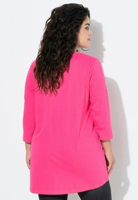 Maglietta rosa a maniche tre quarti in tessuto morbido e leggero. Presenta una silhouette comoda e un orlo arrotondato, abbinata a leggings scuri.