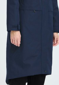 Cappotto impermeabile blu navy con colletto alto, polsini regolabili con Velcro e una texture liscia. Presenta una linea dritta e una chiusura laterale a scatto.