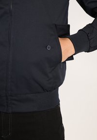 Marinblå bomberjacka med elastiska muddar och midjeband, utrustad med en knappficka och slät tygstruktur.