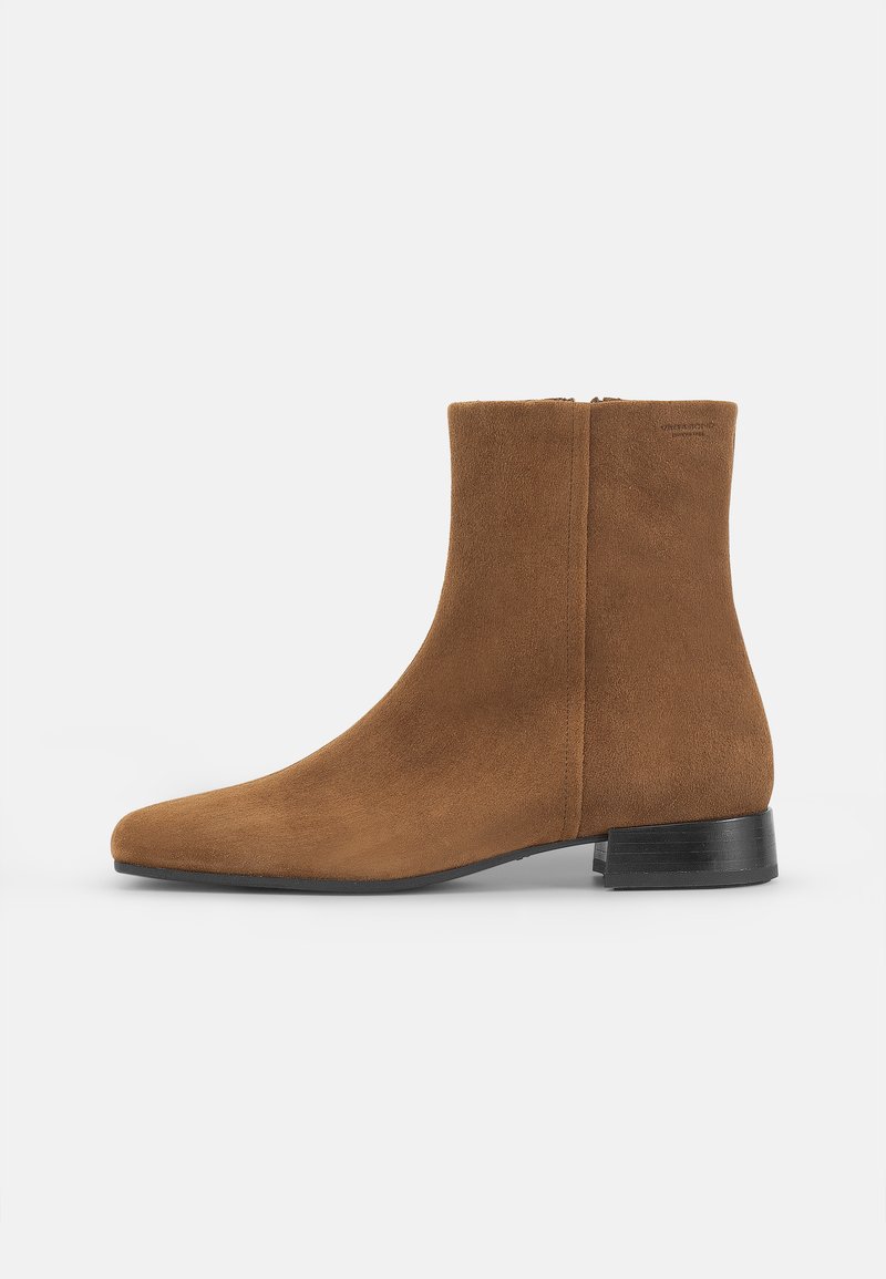 Bota de tobillo de ante marrón con punta afilada, tacón bajo negro y cremallera lateral; textura suave y diseño minimalista sin adornos adicionales.