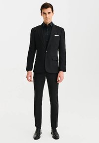 Czarny garnitur o dopasowanym kroju, z pojedynczym zapięciem na guzik i klapami w kształcie notcha. W zestawie z dopasowaną czarną koszulą wizytową i wypastowanymi czarnymi butami.