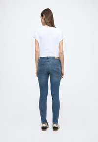 Úzke modré rifľové nohavice s vysokým pásom a štruktúrovanými zadnými vreckami, skombinované s krátkym bielym tričkom s rukávmi. Jemné detaily švov sú viditeľné.