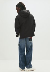 Jachetă neagră cu glugă, cu un croi lejer, asortată cu pantaloni din denim albastru largi, ce au un detaliu grafic roșu și negru pe buzunar.