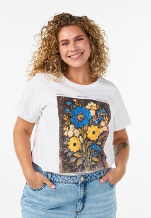 Smilende kvinde med krøllet hår iført hvid t-shirt med blå og gul blomsterprint, hænder i lyseblå jeans lommer.