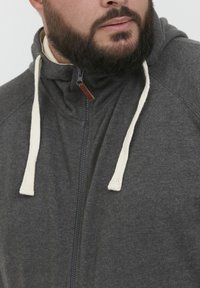 Felpa con zip di colore grigio carbone con cordino color crema. Tessuto morbido, collo alto e zip con un dettaglio in pelle marrone al collo.