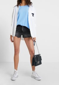 Tenue décontractée composée d'un haut bleu clair, d'une chemise blanche à boutons avec logo, de shorts en denim noirs, d'un sac à main noir et de baskets blanches.