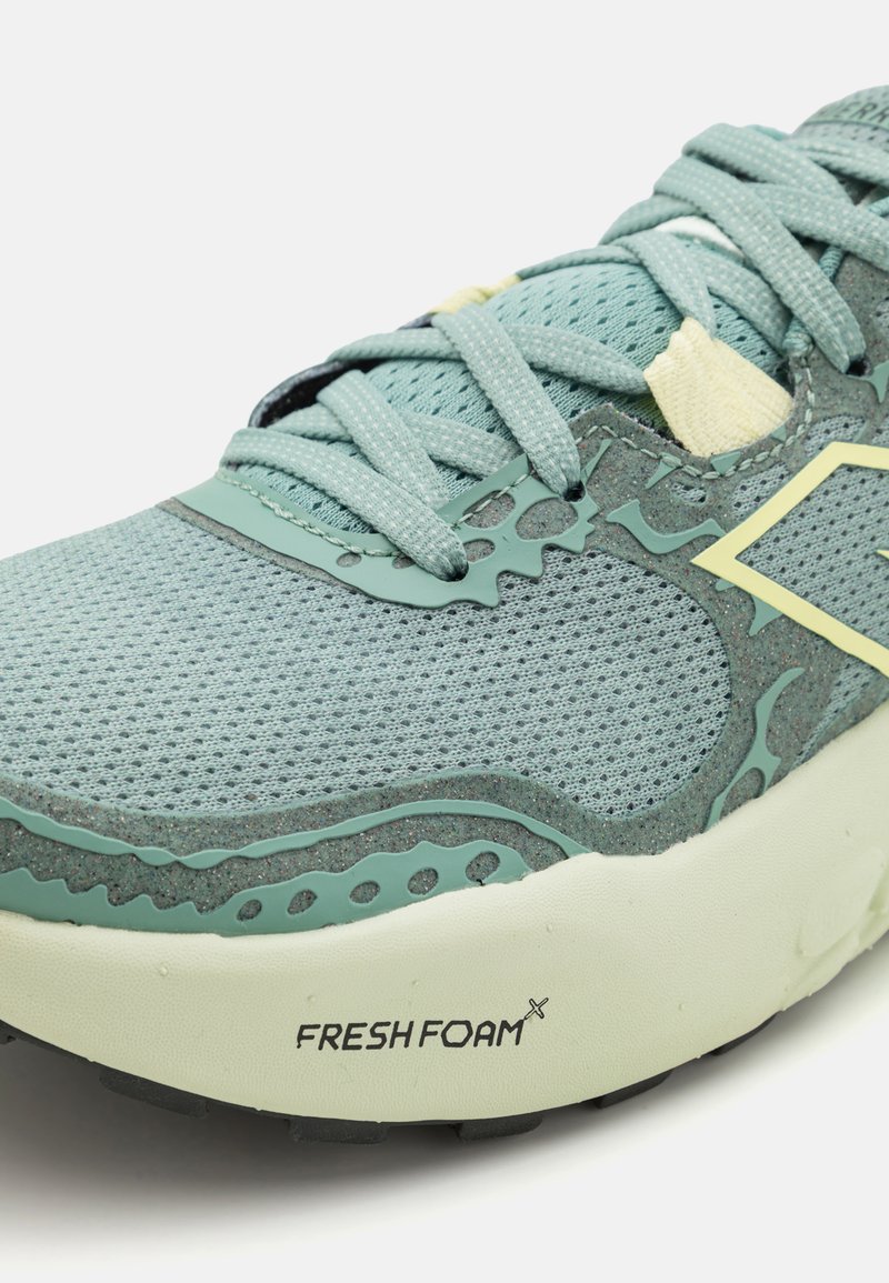 Zapatilla de running de malla en azul claro, con una parte superior texturada, lengüeta acolchada, cordones y marca en la suela. Con detalles en verde.