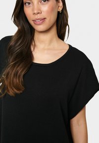 Fekete kötött fels�ő, rövid, széles ujjakkal és kerek nyakkivágással. Simább textúrával és minimális dizájnnal rendelkezik. Arany fülbevalók kiemelve.