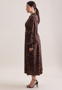 Vestito a stampa leopardata in marrone e nero, maniche lunghe, vita aderente, silhouette fluida, abbinato a sandali neri con tacco alto.