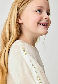 Lys beige sweatshirt med en tekstureret overflade, forsynet med et cremefarvet bånd ned ad ærmet mærket "BAKER" i guldtekst.