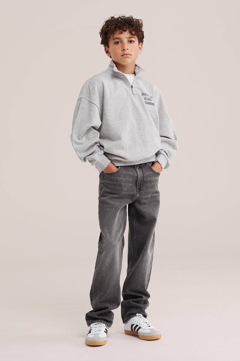Junge mit lockigem Haar steht mit den Händen in den Taschen, trägt ein hellgraues Sweatshirt mit Viertelreißverschluss, dunkle Jeans und weiße Sneakers mit schwarzen Streifen.