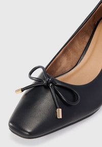 Black leather ballet flat with a square toe, featuring a bow detail with gold hardware accents. The interior is beige suede-like material.