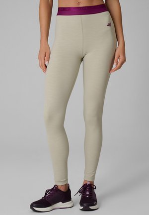 Leggings beige avec une ceinture violette, portés avec des chaussures de sport noires et violettes sur une personne debout sur un fond gris.