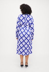 Abito a scacchi blu e bianchi con vita a corsetto, maniche lunghe e colletto. Tessuto morbido con una silhouette fluida. Sandali con tacco neri.
