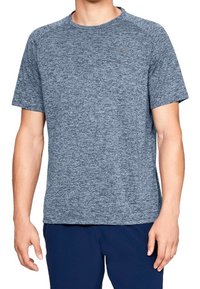 Hombre que lleva una camiseta deportiva de manga corta en gris azulado jaspeado con un pequeño logo en el lado izquierdo del pecho y pantalones azul marino.