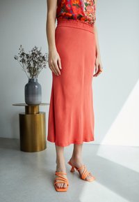 Saia midi coral com cintura elástica, feita de um tecido suave. Combinada com sandálias de salto laranja e uma blusa com padrão floral.