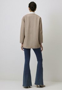 Beżowy, teksturowany, przerośnięty płaszcz z rozcięciem z tyłu, zestawiony z niebieskimi dżinsami o rozszerzanych nogawkach. Strój zawiera białą koszulę z kołnierzykiem pod spodem.