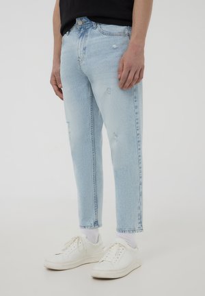 Jeans azzurri chiari con una vestibilità dritta, caratterizzati da toppe strappate e un orlo leggermente accorciato. Abbinati a sneakers bianche e calze semplici.