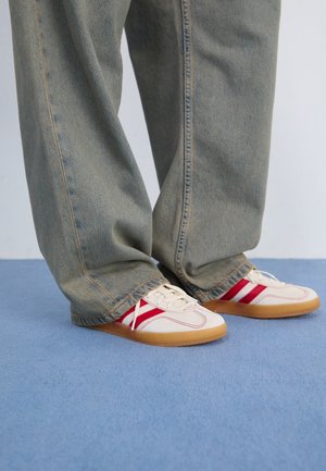 White sneakers with red stripes, textured canvas upper, rounded toe, flat gum sole, paired with loose, faded blue denim pants.