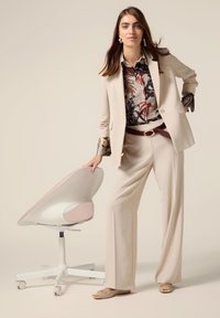 Blazer avorio e pantaloni a gamba larga abbinati a una camicia con motivo. Scarpe beige con dettagli dorati. Sedia da ufficio moderna bianca sullo sfondo.