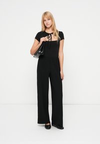 Μαύρη ολόσωμη φόρμα με κοντά μανίκια, τετράγωνη λαιμόκοψη και λεπτομέρεια με κορδόνι. Διαθέτει φαρδιά παντελόνια, ριμπ υφή και συνοδεύεται από μαύρη τσάντα.