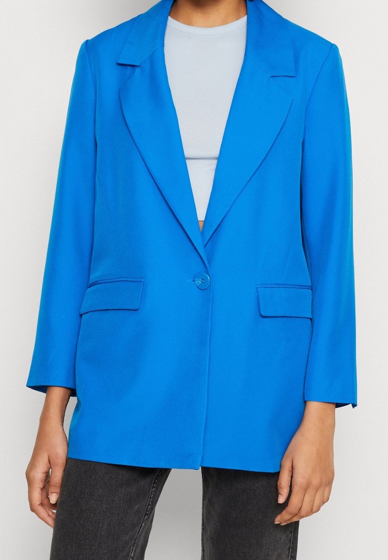 Personne portant un blazer bleu vif à un bouton sur un crop top bleu clair et un jean gris foncé, debout devant un fond blanc.