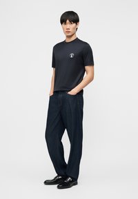 Νεαρός άνδρας στέκεται με τα χέρια στις τσέπες, φορώντας μαύρο κοντομάνικο πουκάμισο με μικρό λευκό λογότυπο και σκούρο φαρδύ τζιν, μαύρα παπούτσια.