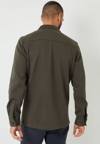 Oliivinvihreä pitkähihaispaita, jossa on terävä kaulus, yksivärinen pinta ja napilliset hihansuut. Selkäpuolella on olkapäiden yli kulkeva pääntien sauma.