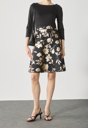 Frau trägt schwarzen Dreiviertelarm-Oberteil und schwarzen Rock mit beigem Blumenmuster, trägt schwarze Schultertasche und schwarze Riemchenabsätze.